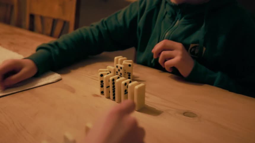 A young boy plays dominoes with another person while seated at the dining table at night