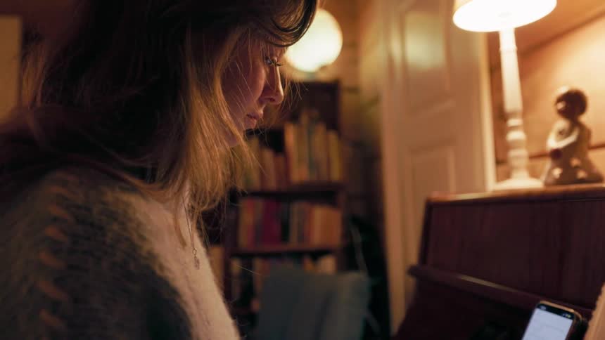 A woman sits indoors looking at her mobile device with a lamp placed on a shelf beside her