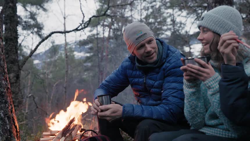 Family at a campsite in the woods next to a campfire drinking and eating from a cup happily