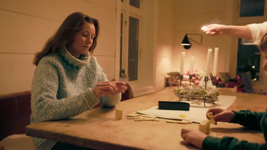 Mother and son playing dominoes at a dinner table at night and next to it a person lighting candles