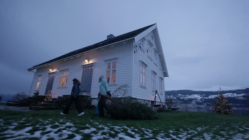 A mother and her two kids all dressed in winter clothing walk toward their home as the mother drags a christmas tree behind her on a cloudy evening