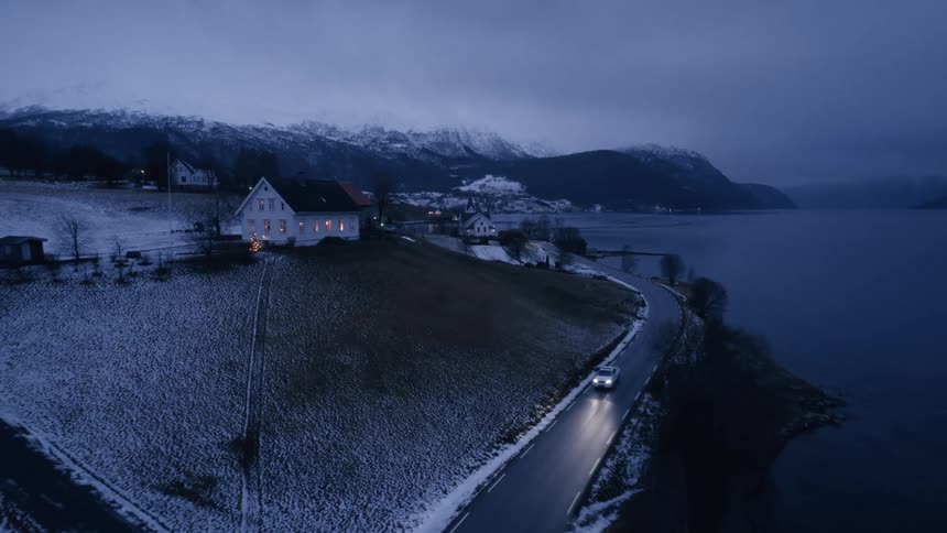 Vehicle with headlights on traveling on a road along a lake with rural houses in the background and snowcovered mountains
