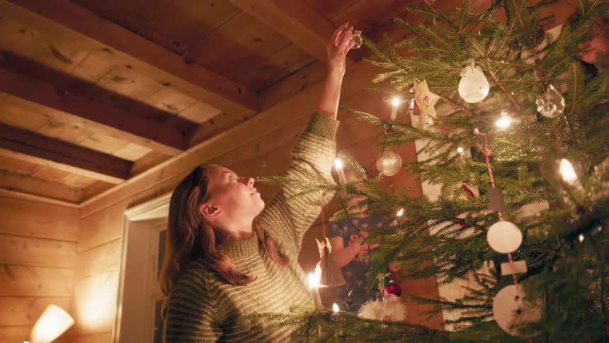 A woman in a sweater reaches up to decorate a lit christmas tree inside a room with wooden walls and a lamp emitting a warm glow