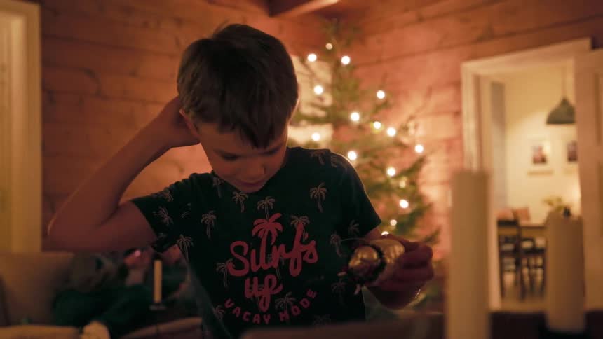 A young boy wearing a palm tree shirt carefully handles christmas decorations in a warmly lit room with a decorated christmas tree standing in the background