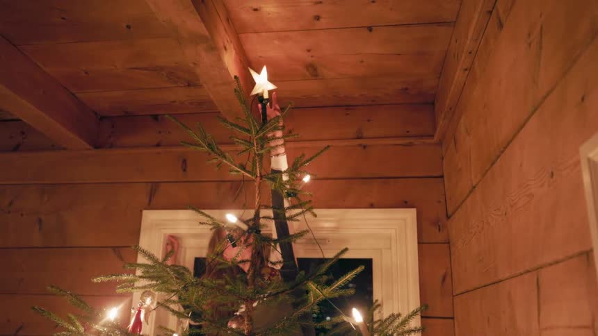 A girl reaches up to place a glowing star on the top of a decorated christmas tree inside a room with wooden walls and ceiling