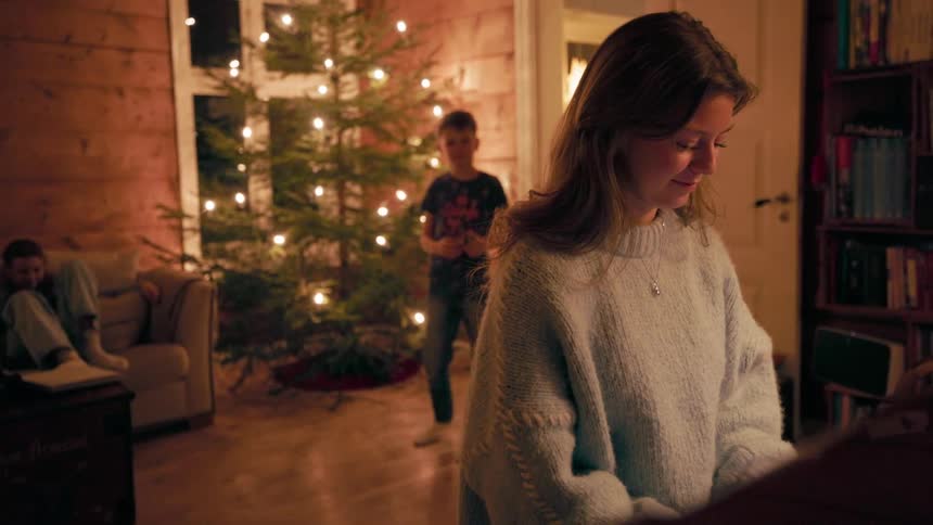 A young mother wearing a sweatshirt plays the piano indoors laughing as a boy moves around the living room behind her near a christmas tree while another child sits on the couch at nighttime