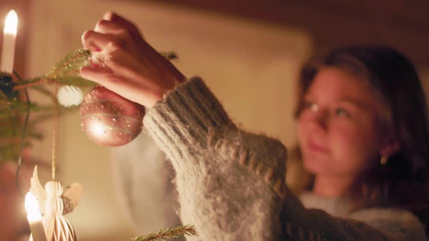 A woman with light brown hair and wearing a lightcolored sweater hangs a shiny ornament on a lit christmas tree