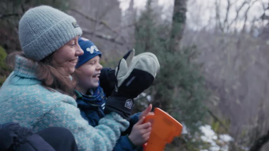 A joyful woman and child sit outdoors in a forested area laughing together and clapping