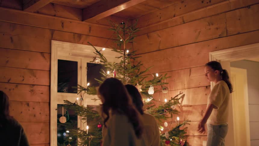 Teenage girls stand admiring a decorated and lit christmas tree inside a rustic wooden room