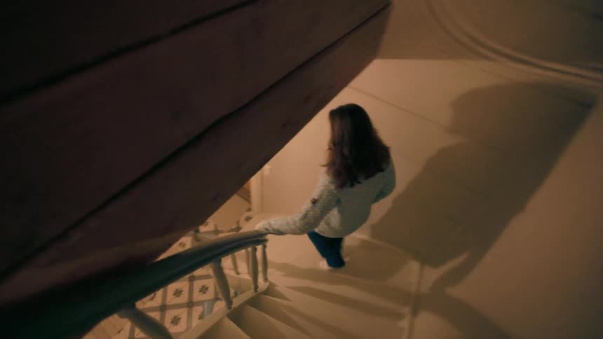 A woman with long hair descends a wooden staircase with a patterned landing in a cozy home illuminated by warm lights