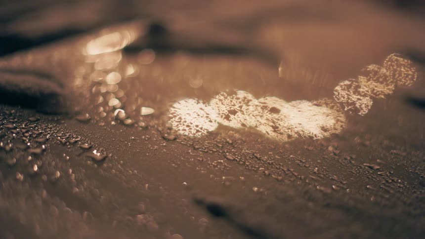 An extreme closeup captures shimmering water droplets and glowing blurred light reflections on a dark textured surface