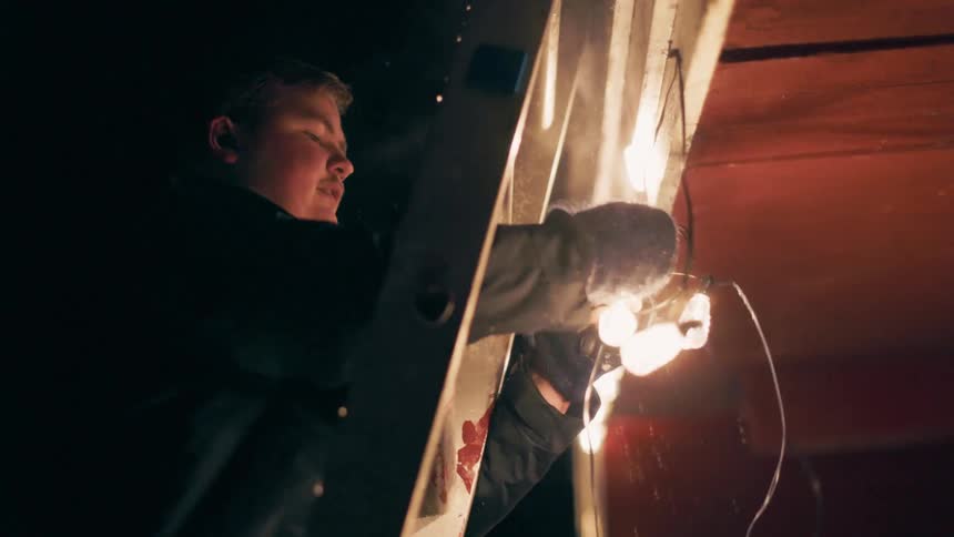 Man on a ladder outside a house at night with snow tying a string with lit decorative lights