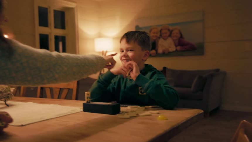 A young boy in pajamas sits at the dining table playing dominoes laughing as his mother playfully pokes his nose at night