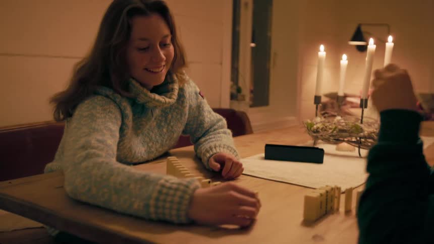 A mother with long brown hair smiles at her son while they arrange dominoes on a wooden table illuminated by candlelight