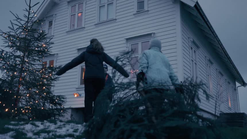 A family carry a large evergreen tree towards a white twostory house at dusk passing by another decorated christmas tree