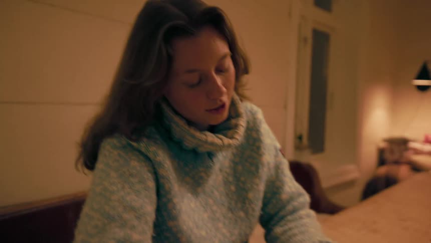 A woman in a blue sweater with long brown hair arranges dominoes on a wooden table illuminated by the gentle glow of warm lamps