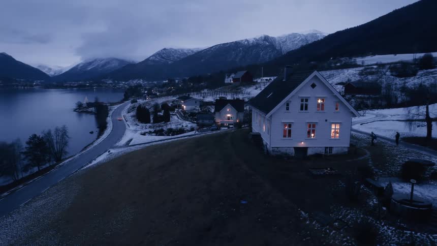 A brightly lit white house nestles on a snowdusted peninsula jutting into a still dark fjord under a cloudy twilight