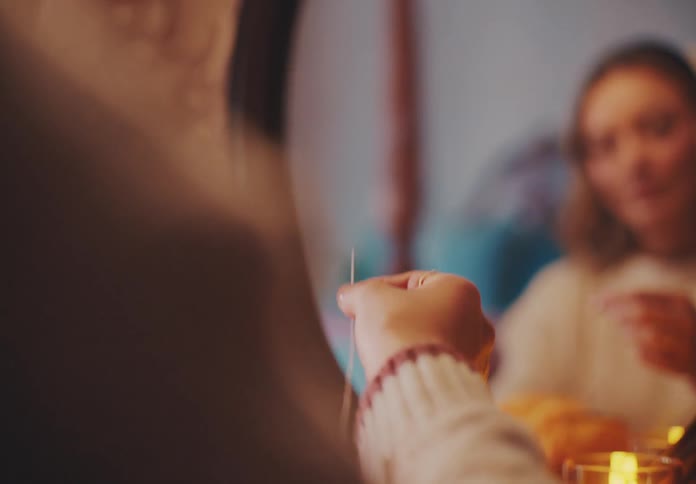 Closeup of a woman holding a needle threaded with thread in front of a mirror with a candle inside a vase nearby in a warmly lit room