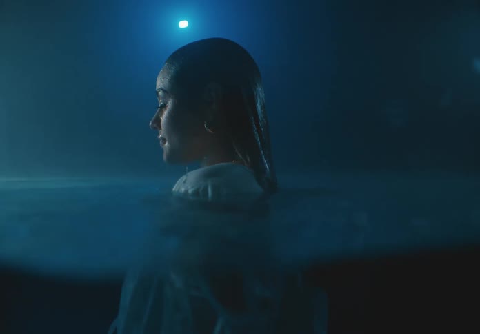 A woman in a white dress floats submerged underwater her head breaking the surface illuminated by a soft dim blue light that envelops the scene