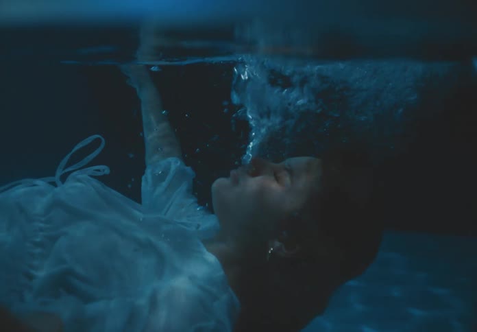 A woman in a white dress floats submerged underwater with bubbles rising from her face