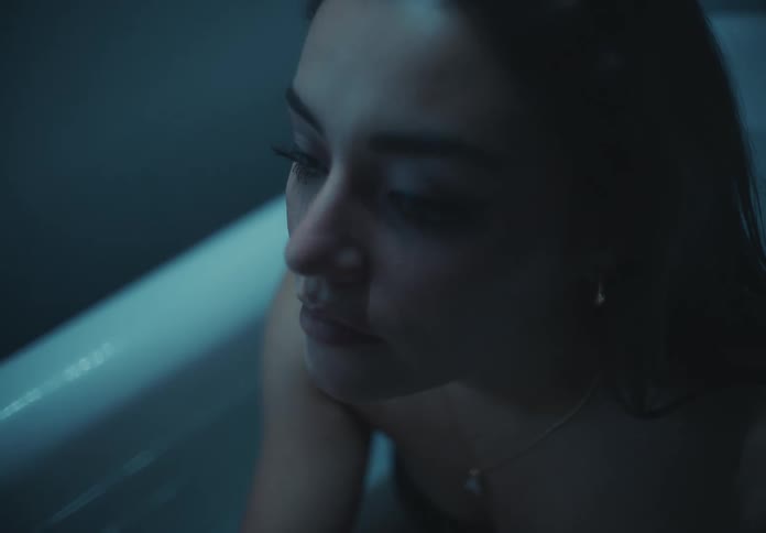 The face of a woman with makeup around her eyes who is sad while sitting in a bathtub with her hands running over her face
