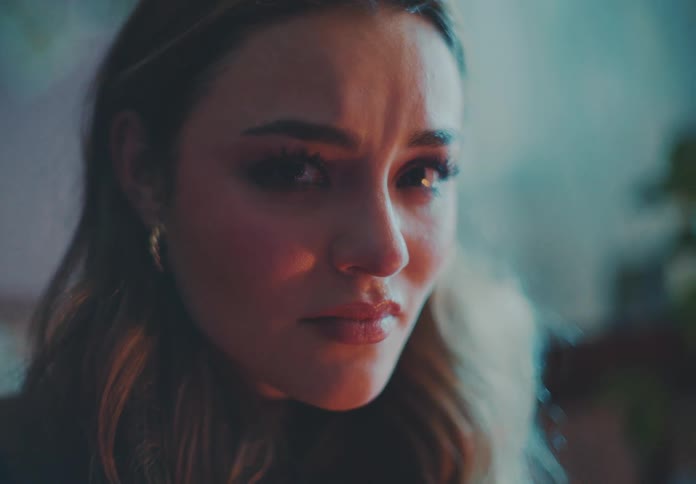 The face of a young woman with makeup around her eyes and wearing gold earrings who is staring straight ahead while having sad tones