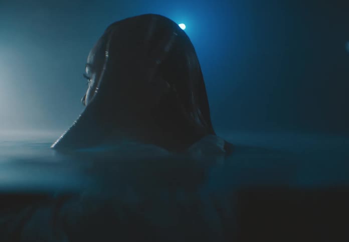 A woman in a white dress floats submerged underwater her head breaking the surface illuminated by a soft dim blue light that envelops the scene
