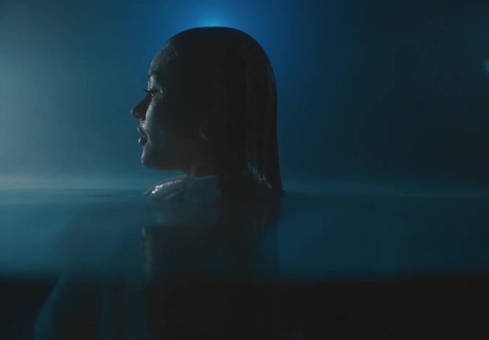 A woman in a white dress floats submerged underwater her head breaking the surface illuminated by a soft dim blue light that envelops the scene