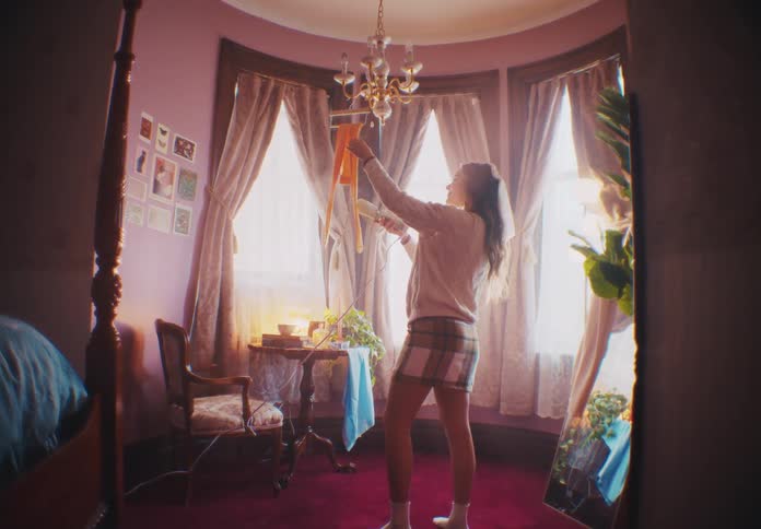 A woman standing in a pink room who is ironing a garment hanging from a chandelier with a hot air iron