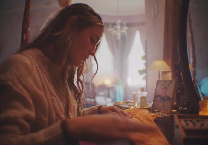 At midday inside a bedroom a woman wearing a sweatshirt examines a yellow tight on a hanger brings it to a table sits down and tries to fix a badge on it