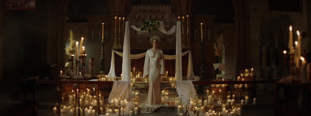 A woman in a white gown and ornate headpiece stands quietly at the center of a candlelit altar in a dimly lit church