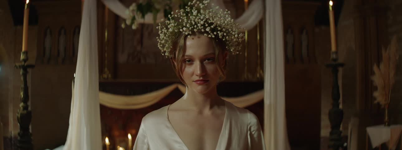 A young woman in a white robe and babys breath crown stands centered in soft ceremonial light gazing into the camera