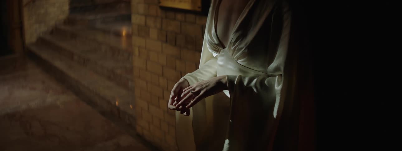 A woman in a silky flowing garment stands near dimly lit stone steps gently holding her own hands