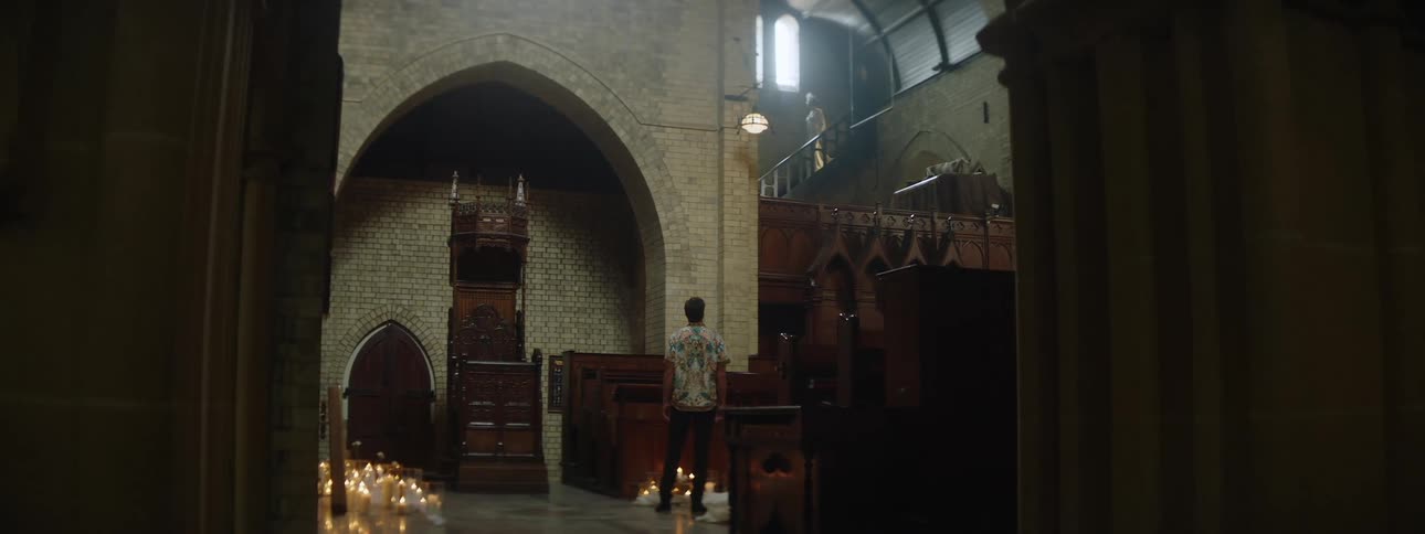 A man stands alone inside a large dimly lit church or cathedral gazing upward toward a person on the balcony