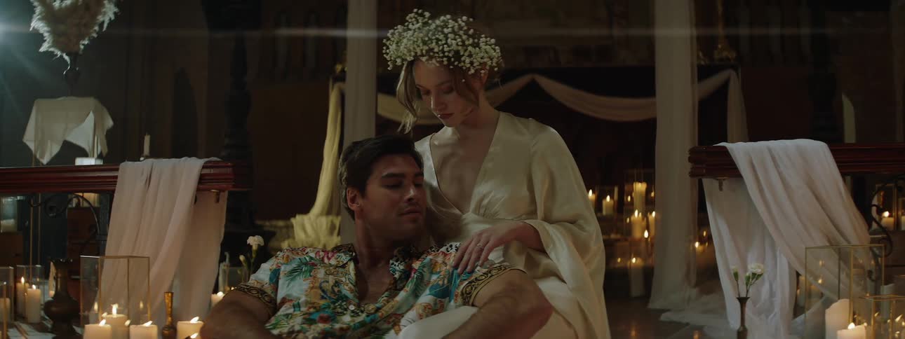 Seated man in a patterned shirt is touched on the shoulder by a woman in a light dress and a flower crown in a room decorated with candles and white fabrics
