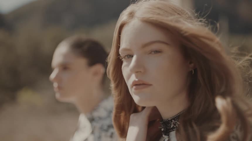 A young woman looks into the camera with another person blurred in the background outdoors