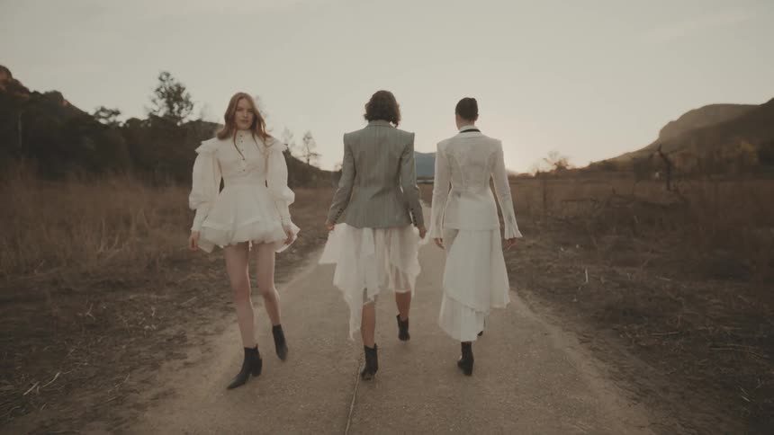 Three stylish women in fashionable attire walk away on a dirt path in a field against a scenic mountain backdrop