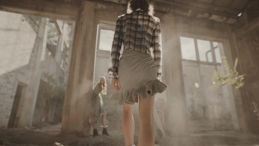 A woman in a plaid top and light skirt walks away from the viewer in a dusty dilapidated building with a blonde woman in the background