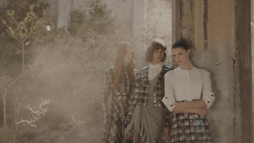 Three fashion models pose in highfashion outfits beside a weathered concrete wall in a dry overgrown setting