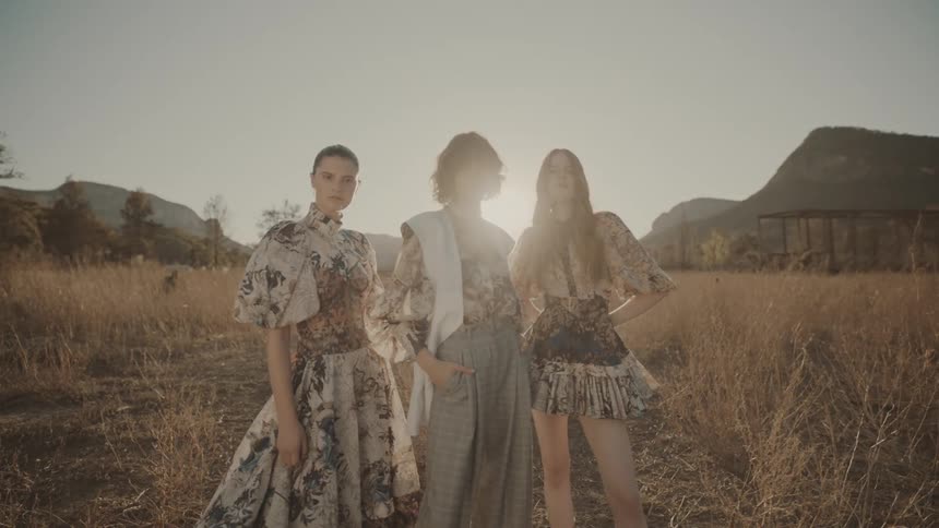 Three women wearing haute couture outfits with floral prints pose in a dry field at dusk with mountains in the background