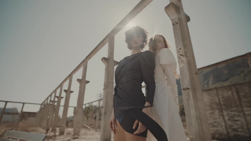 Two women one wearing a black dress and the other wearing a white dress pose in an unfinished structure