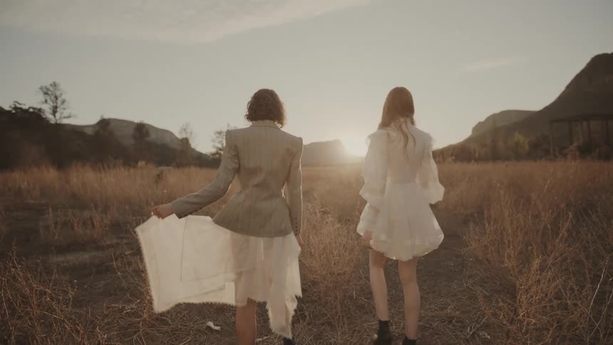 Two young teenage girls on a grassland at sunset both wearing white dresses with one also wearing a suit spinning in a full 360degree turn