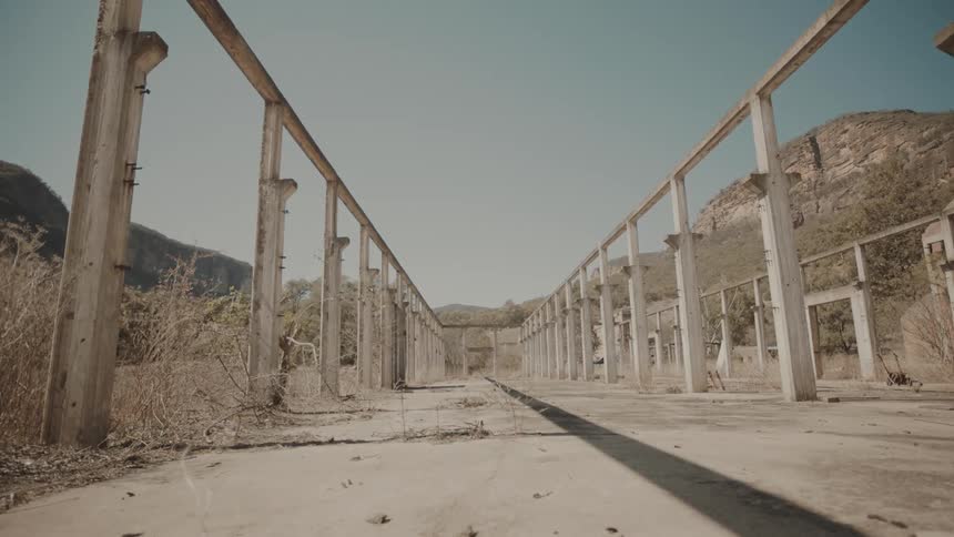 An abandoned structure with concrete pillars and beams surrounded by dry vegetation and hills under a clear sky