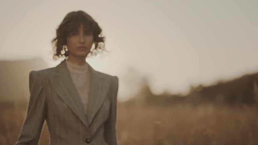 Woman in blazer walking in a field with reeds at sunset