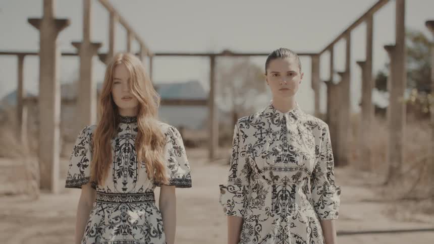 Two models wearing intricately patterned dresses walk side by side in an abandoned industriallooking area with concrete pillars and beams