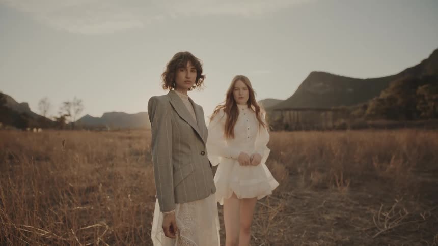 Two models are standing in a dry grassy field with mountains in the background one wearing a gray blazer over a white dress and the other in a white ruffled dress