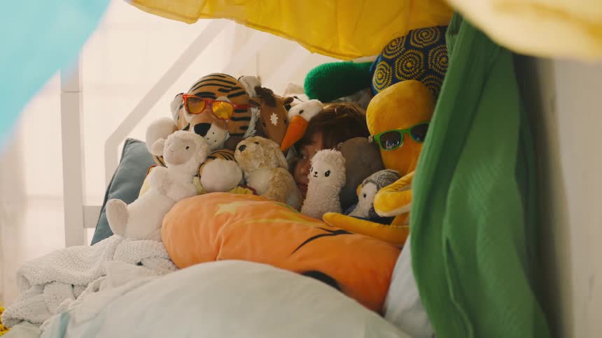 A child is cozily nestled among a pile of stuffed animals and pillows inside a blanket fort