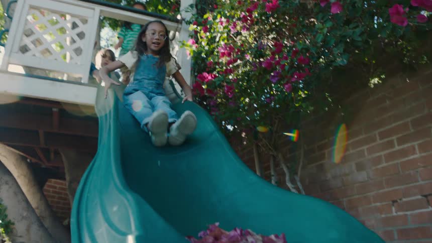 Three children joyfully go down a slide in a backyard play area surrounded by flowers