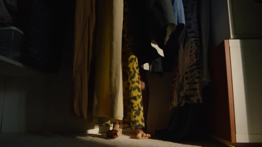A child is hiding barefoot in a dimly lit closet behind hanging clothes
