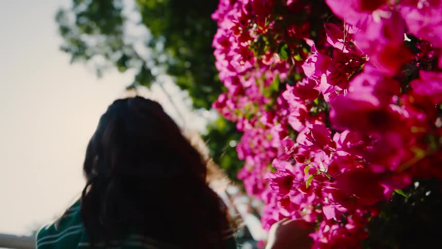 A girl playfully closes her eyes while standing next to vibrant pink flowers in warm sunlight